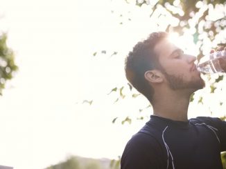 Side view of a young man drinking water from a plastic bottle outdoors in sunlight.
