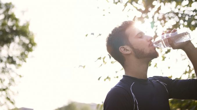Side view of a young man drinking water from a plastic bottle outdoors in sunlight.